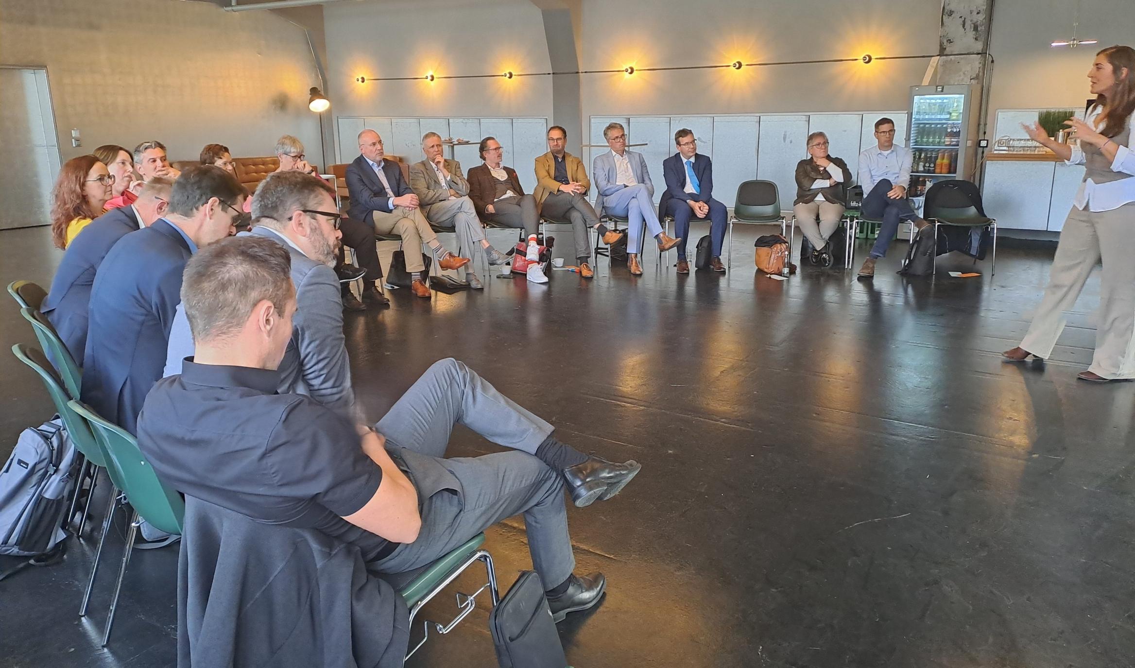 viele Personen sitzen im Halbkreis in einem Tagungsraum und hören einer Sprecherin zu.