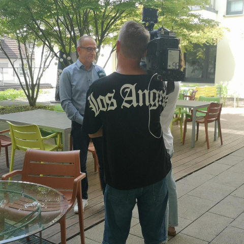 Kameramann und Reporterin im Innenhof mit Vorstand vor Bäumen