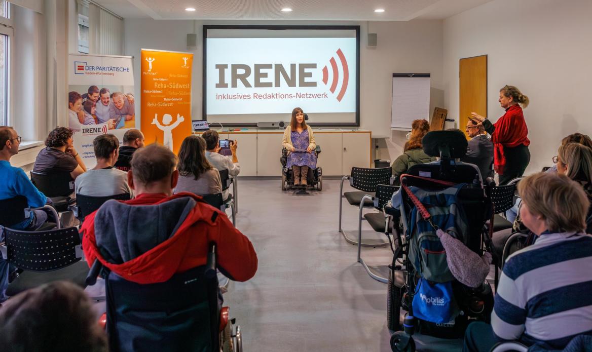 Teilnehmer*innen sitzen im Saal. Frau Nora Welsch sitzt vorne im Rollstuhl und stellt sich vor.