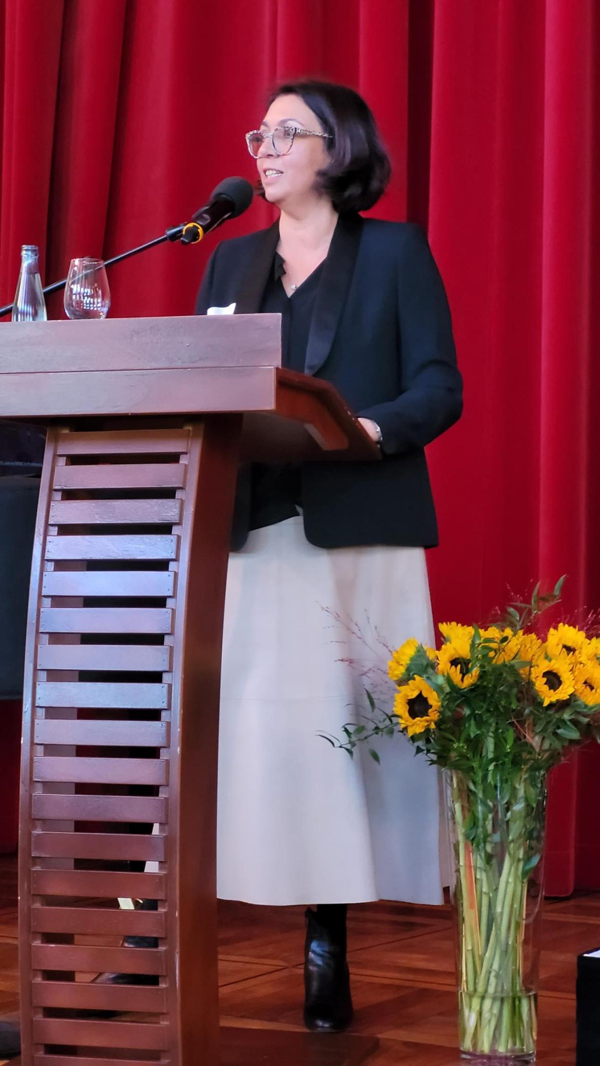 Eine Frau steht am Rednerpult und spricht ins Mikrophon, neben ihr stehen Blumen