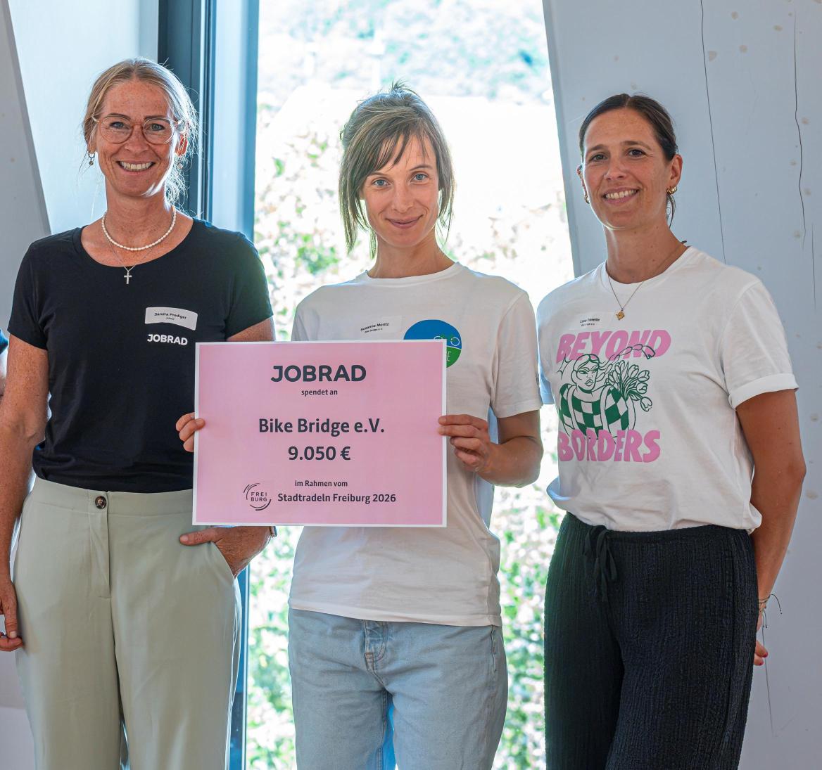 Drei Frauen stehen zusammen, die mittlere hält ein Plakat in der Hand auf dem steht, dass Jobrad 9050 Euro an Bikebridge spendet