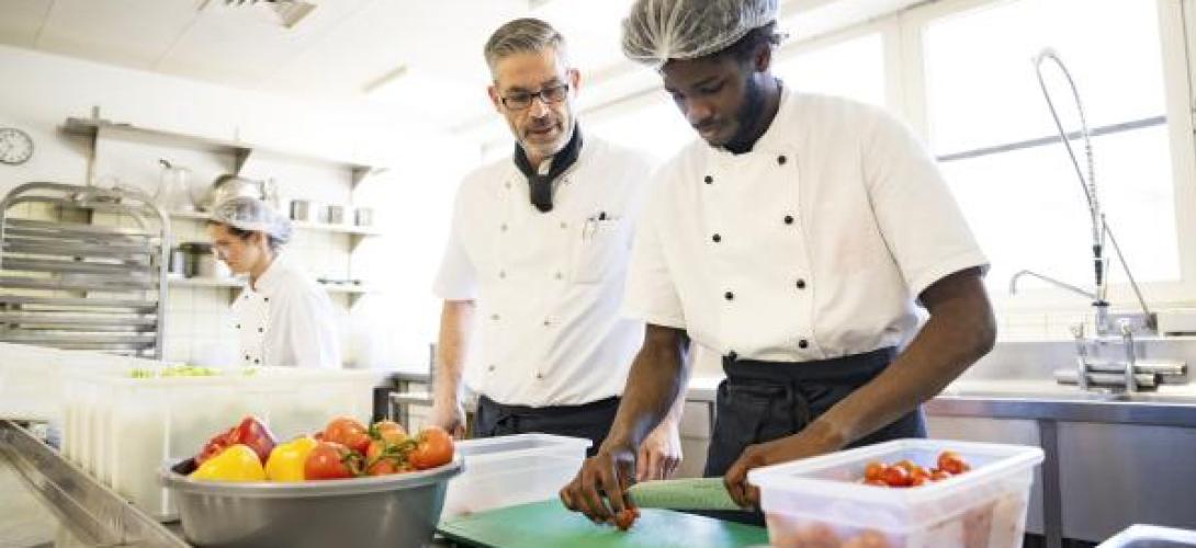 ein junger Mann in Koch-Outfit legt eine Erdbeere auf ein Backblech, ein Anleiter steht neben ihm