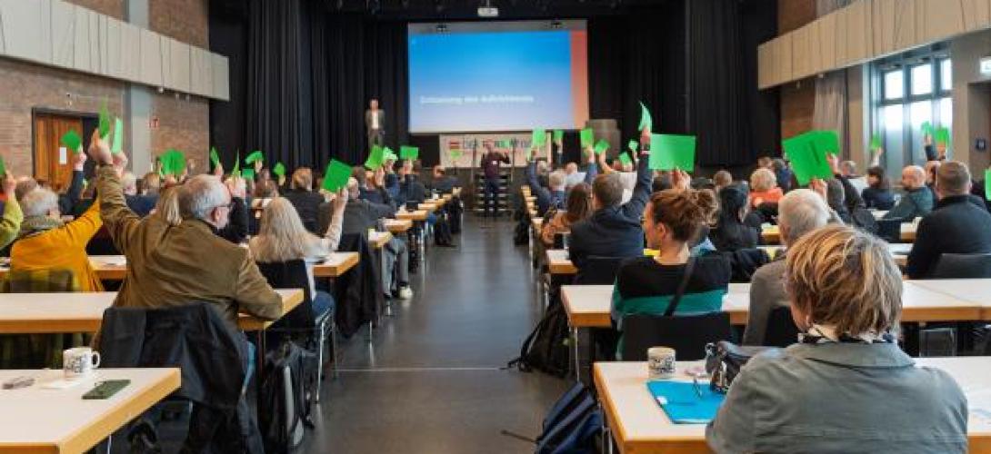 Ein Tagungssaal mit Blick auf eine Bühne, die Tagenden heben grüne Stimmzettel in die Höhe. 