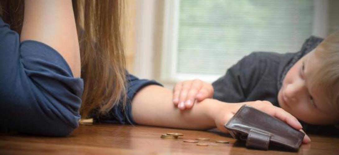 Ein Kind hat seinen Kopf auf den Tisch gelegt und schaut seine Mutter an, die ratlos in ihre Geldb&ouml;rse schaut