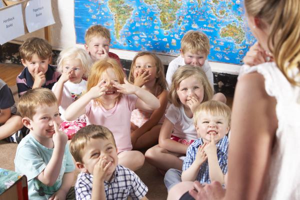 Kleine Kinder deuten gemeinsam mit junger Frau mit Hand auf Mund, Nase oder Stirn