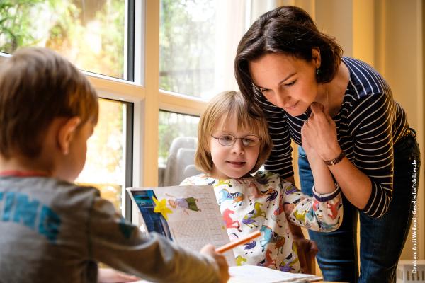 Zwei Kinder am Tisch machen Hausaufgaben. Mutter sieht sich die Aufgaben an.
