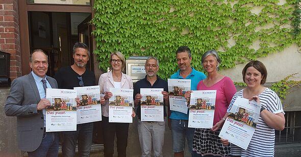 ier M&auml;nner und dreu Frauen stehen nebeneinander im Halbkreis vor einer mit Efeu bewachsenen Hauswand und halten das Kampagnenplakt hoch mit der Aufschrift "Das passt. Kindern Familie erm&ouml;glichen".