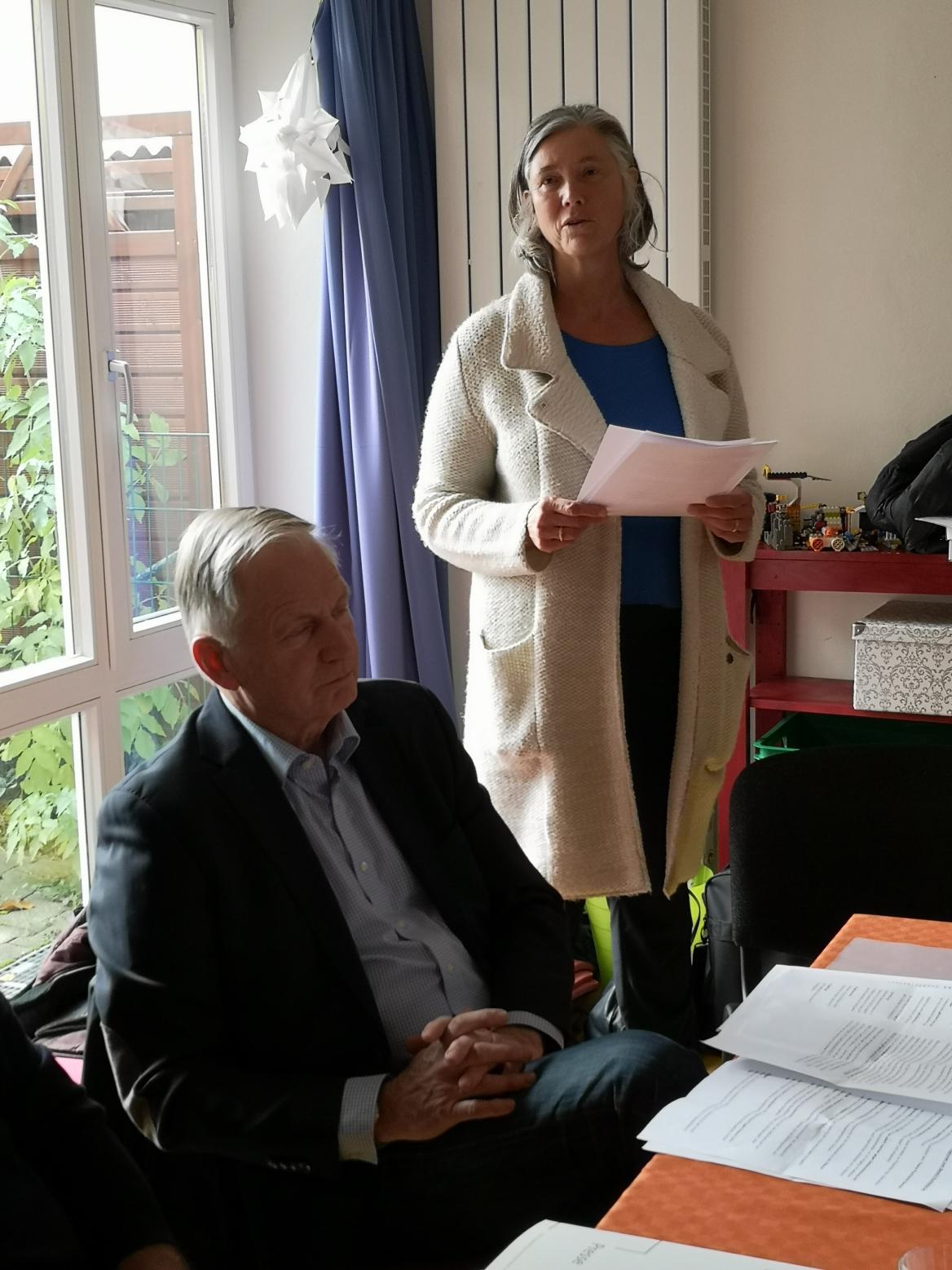 Sitzender Mann in dunklem Anzug und stehende Frau mit wei&szlig;er langer Jacke